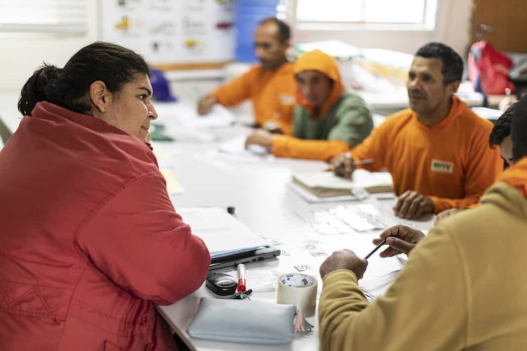 mesa de sala de aula com professora de jaqueta vermelha e quatro trabalhadores da MRV do projeto Escola Nota 10